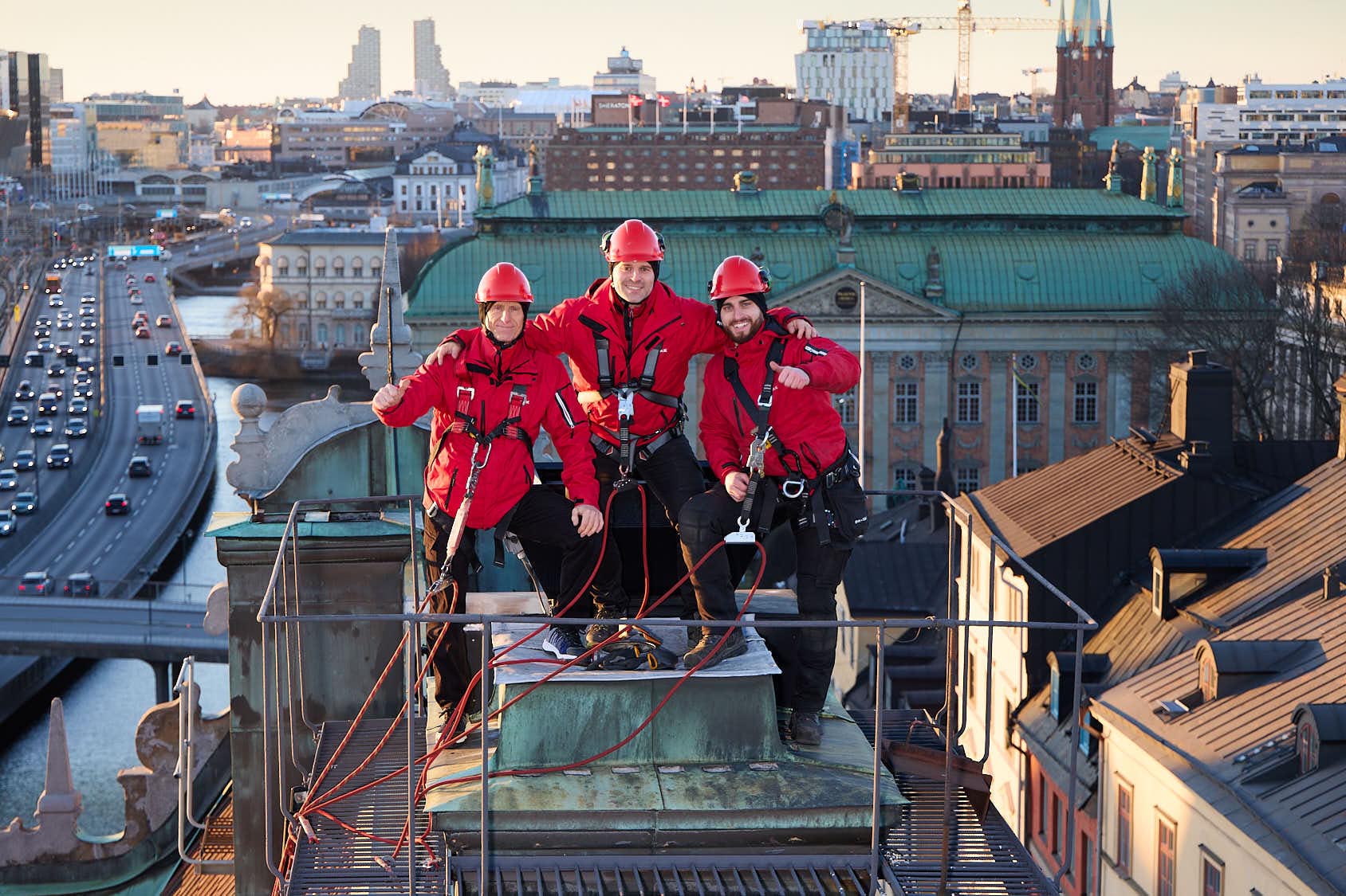 Generationsfoto: tre takarbetera tillsammans i röda arbetsjackor och sele står på skorsten med stadsvy, fotograferat ovanifrån. Foto Martin Ekwall.