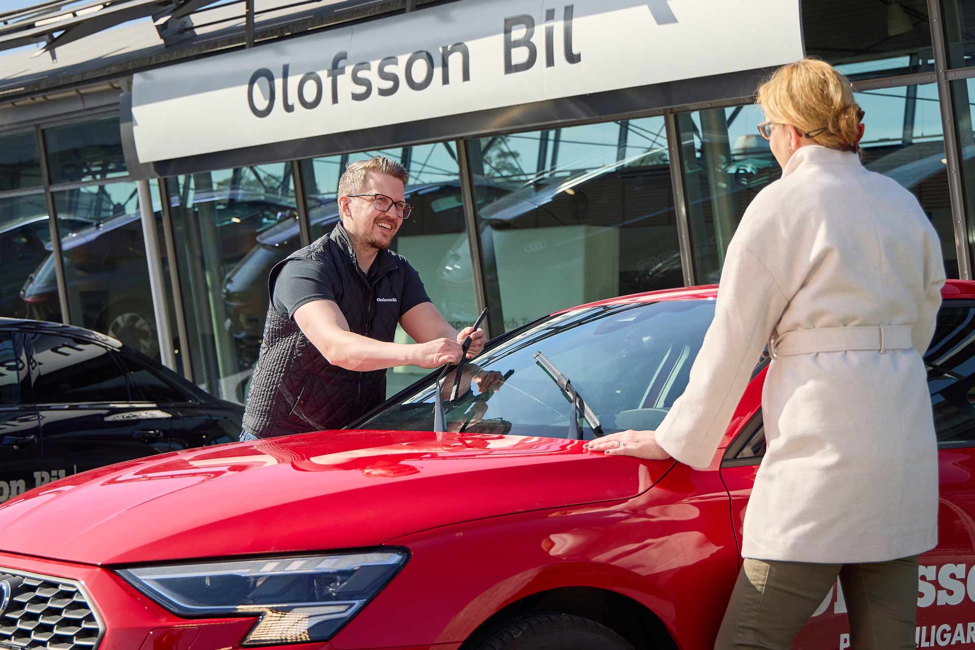 Personal hos Olofsson Bil byter torkarblad på röd bil och hjälper kund utanför bilhallen. Foto Martin Ekwall.