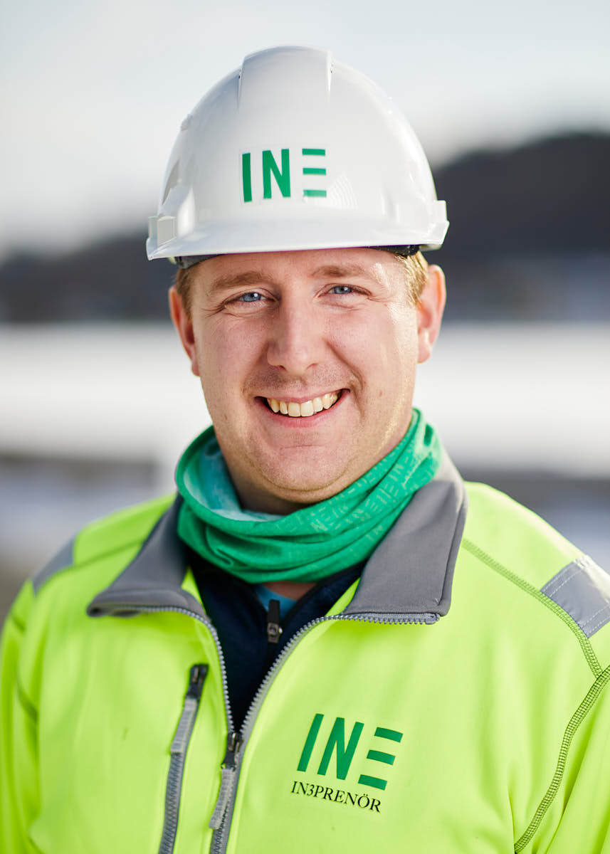 Porträtt av manlig medarbetare på Intreprenör, leendes med grön halsduk i byggkläder och hjälm, utomhus på byggarbetsplats Uppsala.