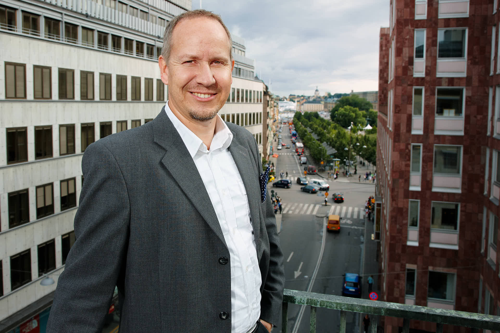 Man leende i grå kavaj står på balkong med utsikt över Stockholm Kungsträdgården, porträtt för Svenska Möten kundcase. Foto av Martin Ekwall.