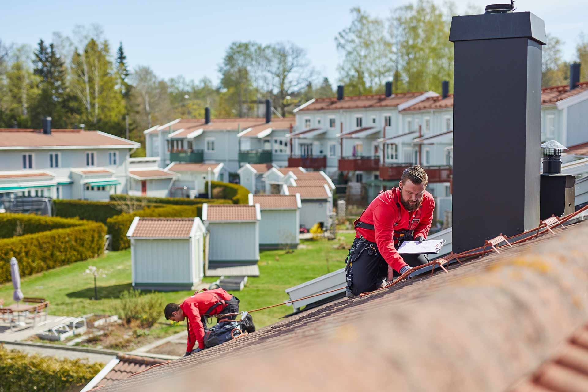 DM TAK takservice. Två takarbetare utför takservice på brant tak med skorsten i förgrunden och villakvarter i bakgrunden. Foto Martin Ekwall.
