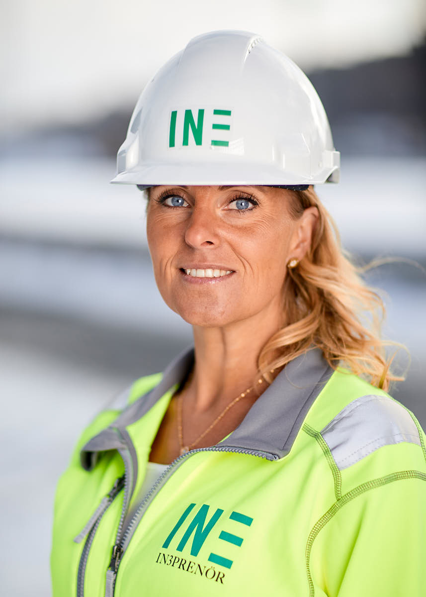 Porträtt av solbränd kvinnlig leendes medarbetare på Intreprenör, byggkläder och hjälm, utomhus på byggarbetsplats Uppsala.