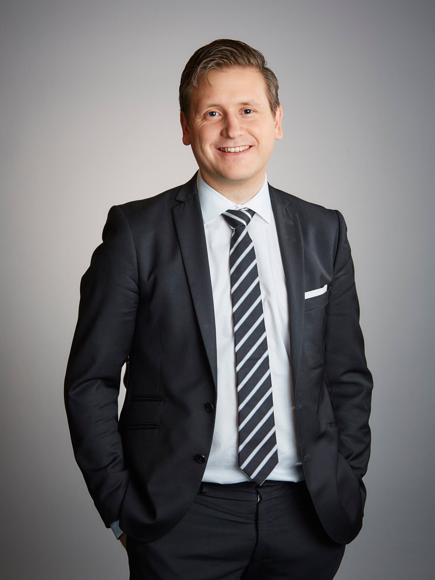 Ramirent styreslemedlem, porträtt i halvfigur av leende man i svart kostym obåda handerna i byxfickorna, ledig pose mot grå bakgrund. Foto Martin Ekwall.