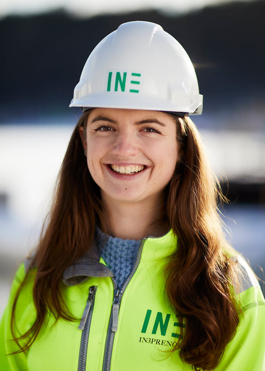 Porträtt av ung glad brunett som är medarbetare på Intreprenör, byggkläder och hjälm, utomhus på byggarbetsplats Uppsala.
