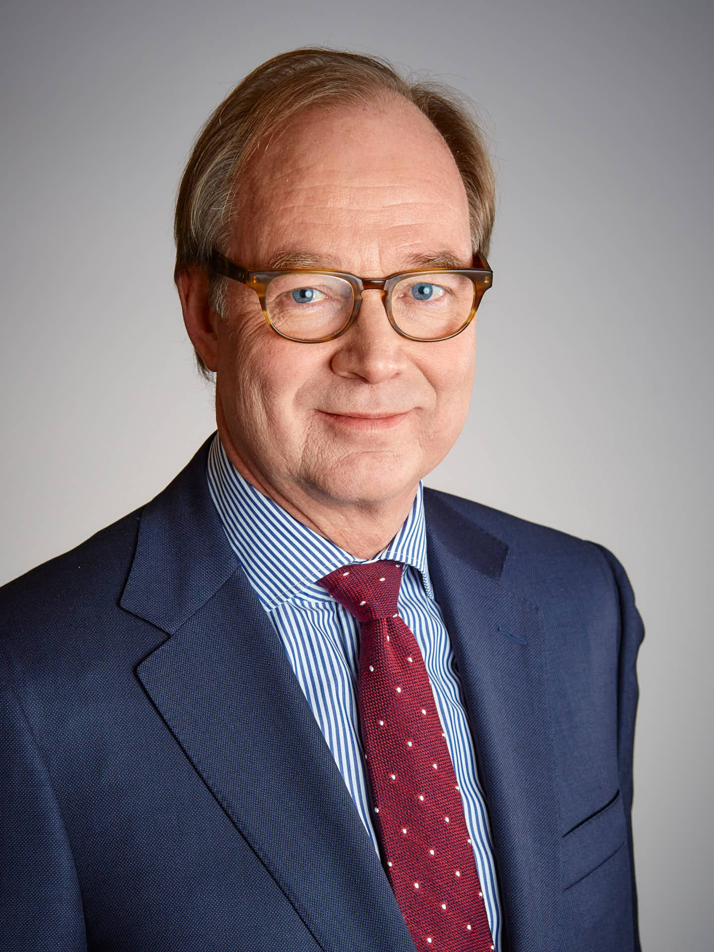 Ansiktsporträtt av man i mörk kostym med blå-rutig skjorta och röd-prickig slips mot grå bakgrund, Ramirents styrelse. Foto Martin Ekwall
