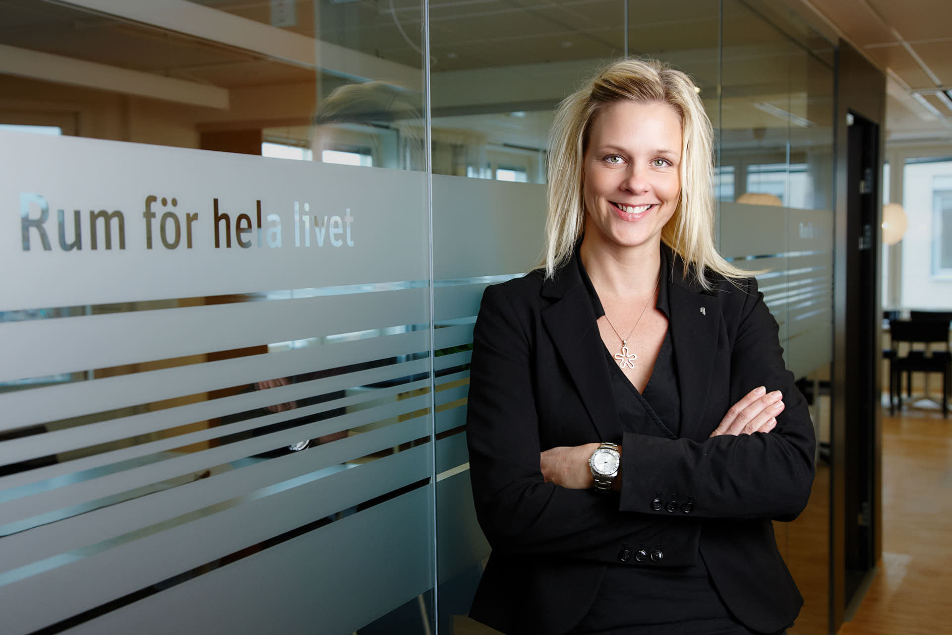 Leende kvinna i svart kavaj står med armarna i kors framför glasvägg med texten “Rum för hela livet”. Kundcase för Svernska Möten.  Foto Martin Ekwall.