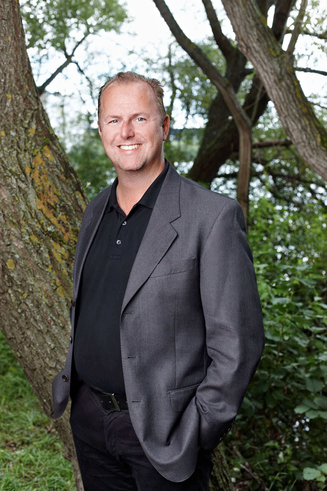 Porträtt av tidigare medarbetare Johan Rosenlind från 3L Systems. Leende i kavaj med händerna i fickorna framför trädstam. Foto av Martin Ekwall