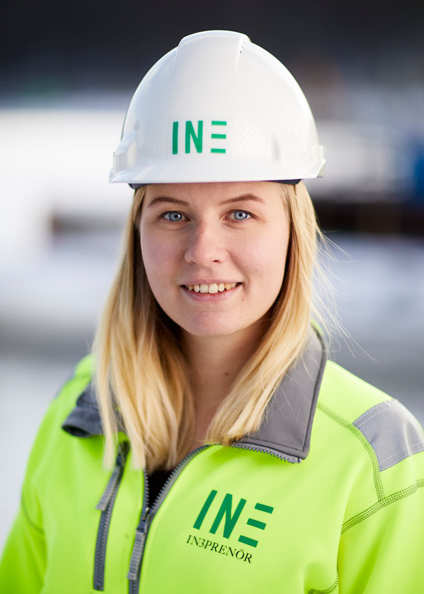Porträtt av ung leendes blond kvinna medarbetare på Intreprenör, byggkläder och hjälm, utomhus på byggarbetsplats Uppsala.