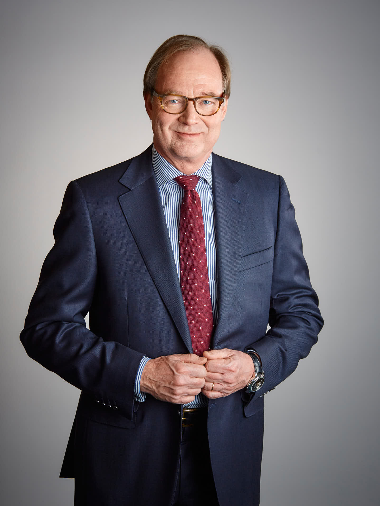 Ramirent styreslemedlem, porträtt i halvfigur av man i blå kostym, röd slips med vita prickar och glasögon, håller händerna i kvajen mot grå bakgrund. Foto Martin Ekwall.