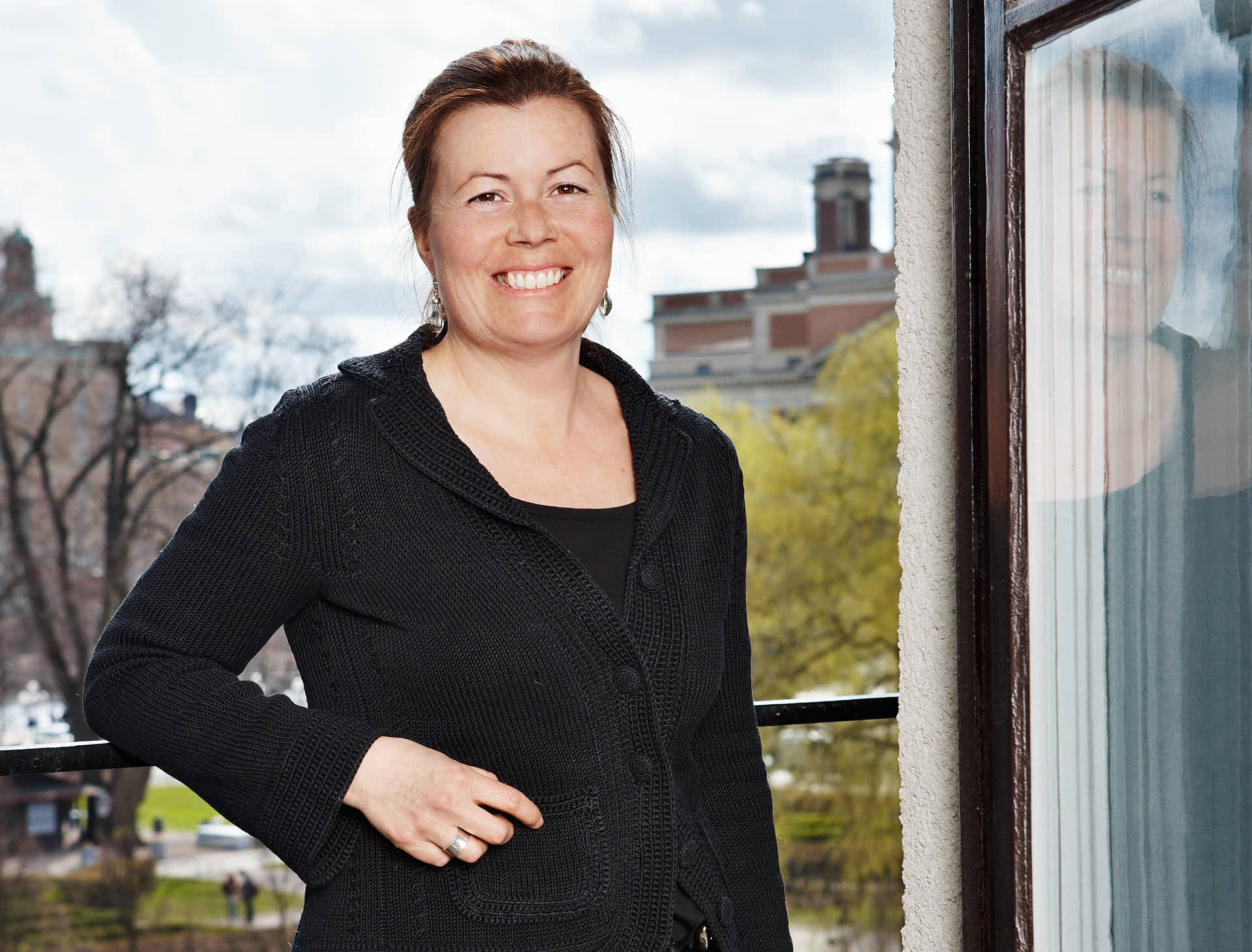 Leende kvinna står på balkong med stadsutsikt i bakgrunden, solig dag med stadens byggnader synliga. Kundcase för Svenska Möten. Foto Martin Ekwall.