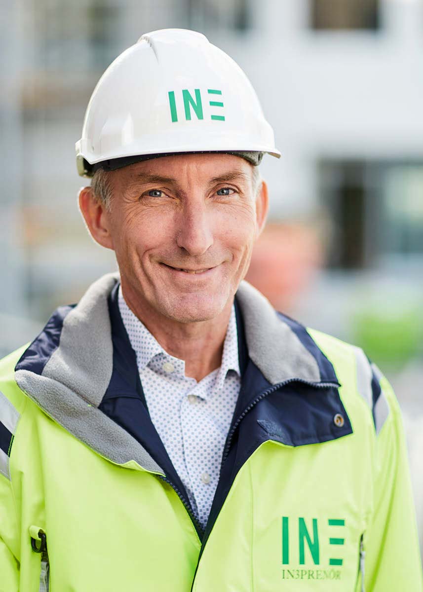 Porträtt av manlig leendes medarbetare medelålders på Intreprenör, byggkläder och hjälm, utomhus på byggarbetsplats Uppsala.