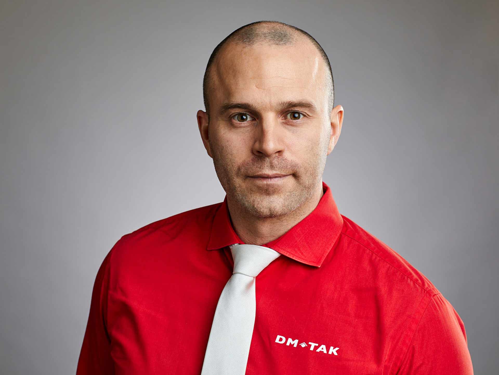 Porträtt av VD David Frykman i röd DM-Tak-skjorta mot ljus bakgrund, mer seriöst uttryck. Foto Martin Ekwall.