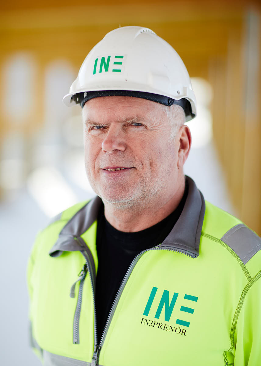 Porträtt av äldre manlig medarbetare på Intreprenör, byggkläder och hjälm, utomhus på byggarbetsplats, idrottsarena i Uppsala.
