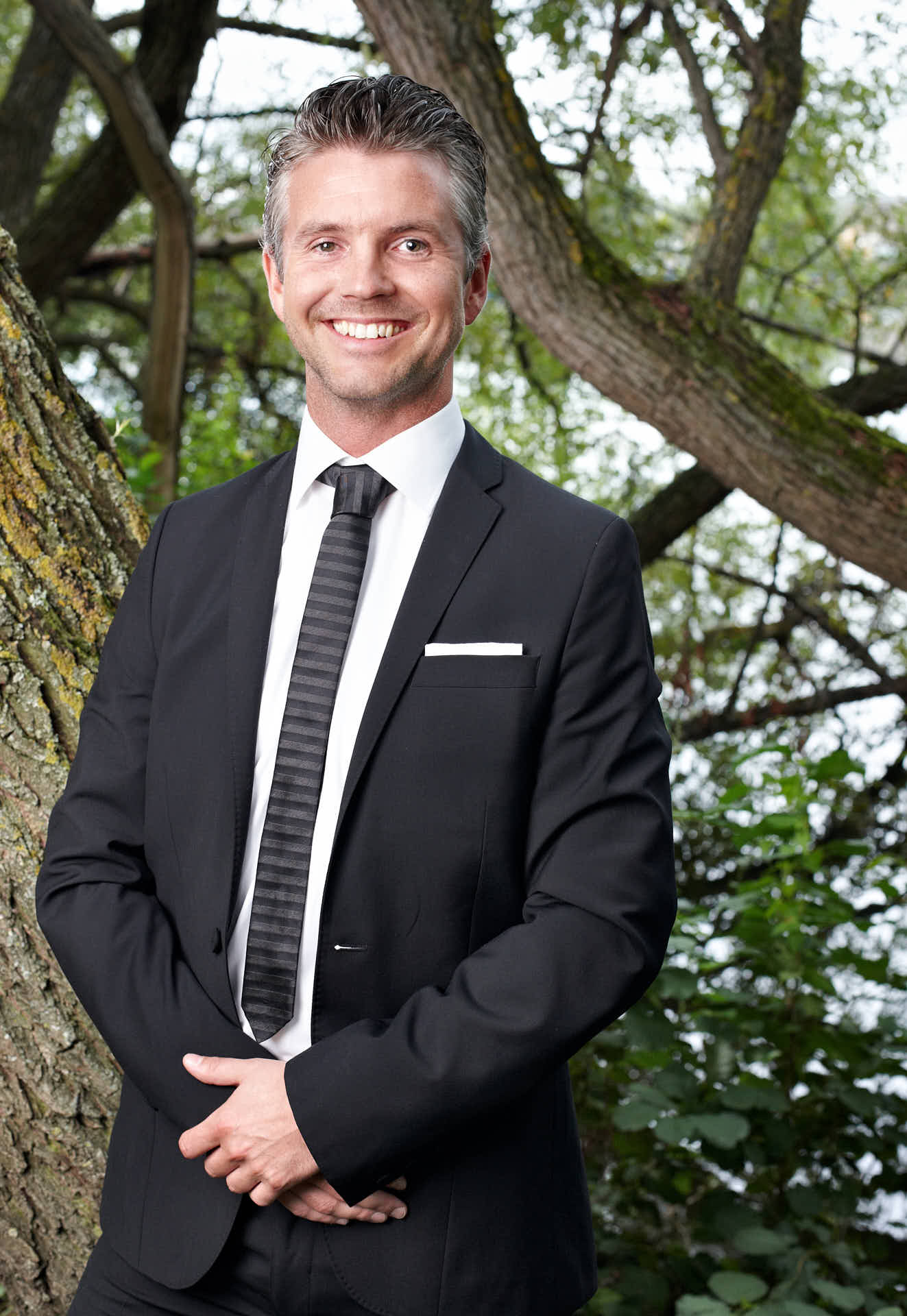 Porträtt av tidigare medarbetare Marcus Bernhardsson från 3L Systems. Leende i kostym vid träd. Foto av Martin Ekwall.