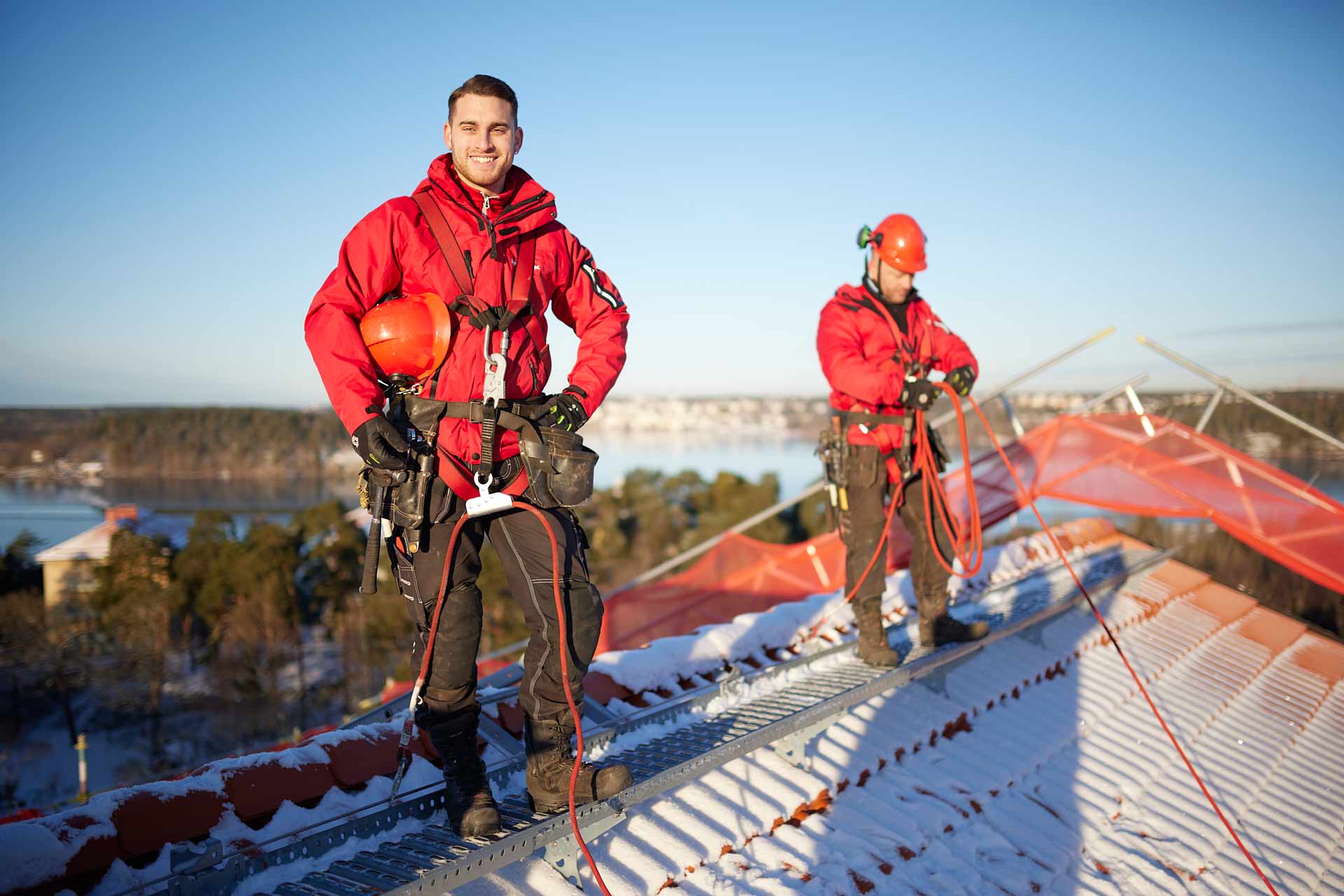 Porträtt av takarbetare i sele på snötäckt tak med utsikt över vatten och vinterlandskap. Foto Martin Ekwall.