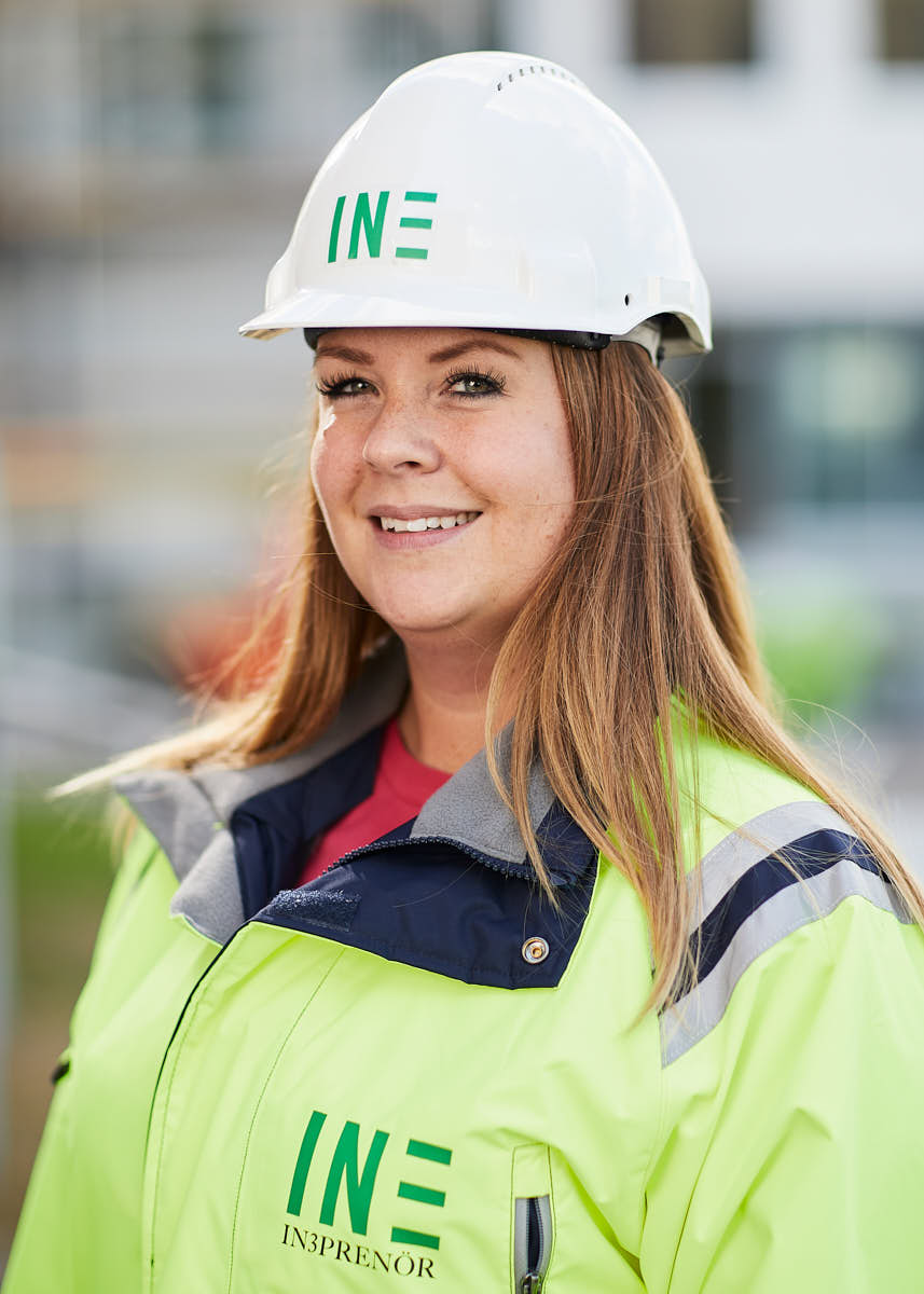 Porträtt av ung leendes kvinnlig brunett medarbetare på Intreprenör, byggkläder och hjälm, utomhus på byggarbetsplats Uppsala.
